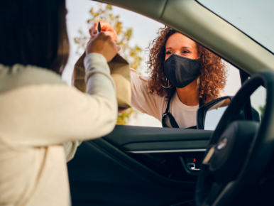 Picture of a woman wearing a mask handing a bag to a person through the window of a car