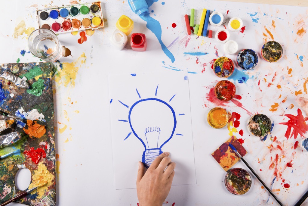 Picture of a hand drawing a lightbulb with paints and other art supplies around the image